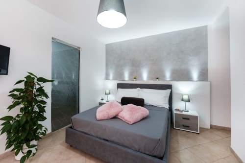 Modern double bedroom at Origenes Rooms, Procida with gray bed, pink pillows, white headboard, green plant, and ceiling lamp