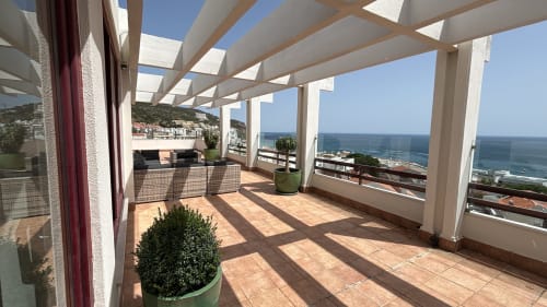 Panoramic penthouse terrace in Sesimbra with sea views, white pergola, potted plants, and coastal landscape beyond railing