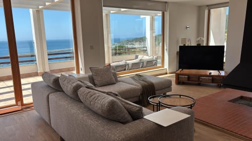 Panoramic penthouse living room with gray sectional sofa, flat-screen TV, and expansive sea views of Sesimbra coast through