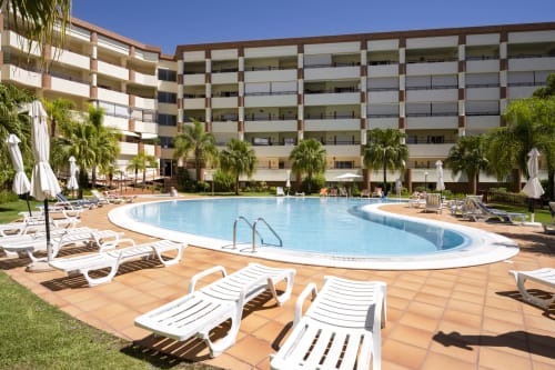 Área de piscina do RB Algarve com espreguiçadeiras, palmeiras e moderno edifício de apartamentos em Vilamoura, Portugal
