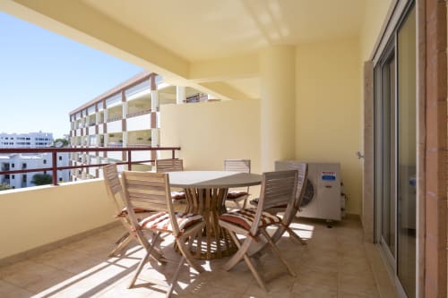 Varanda ensolarada do apartamento RB Algarve Vilamoura com mesa de jantar, cadeiras, ar-condicionado e vista para piscina