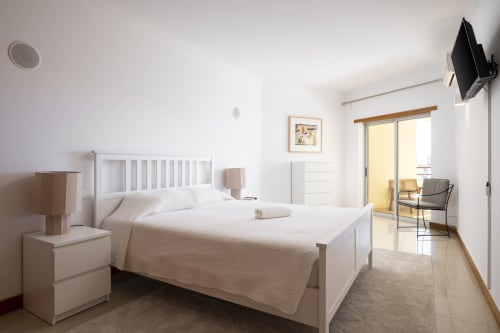 Quarto moderno do apartamento RB Algarve Vilamoura com cama branca, criado, TV de tela plana e portas de vidro para varanda