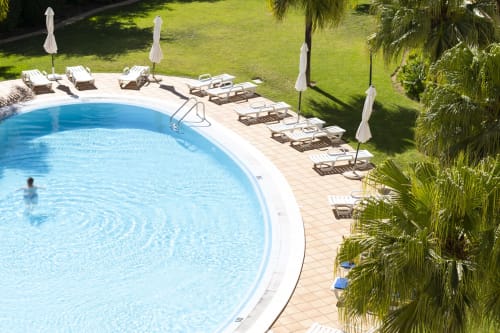 Piscina curvilínea com água cristalina azul no hotel RB Algarve em Vilamoura, Portugal, cercada por espreguiçadeiras, guarda-sóis e vegetação exuberante