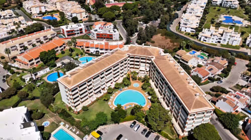 Vista aérea do complexo de apartamentos RB Algarve em Vilamoura com edifícios curvos, piscina central, áreas verdes e arredores