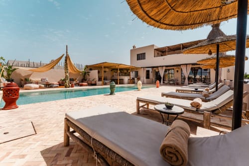 Espace de détente au bord de la piscine du Riad Arkaya à Essaouira avec transats, parasols de paille et piscine turquoise