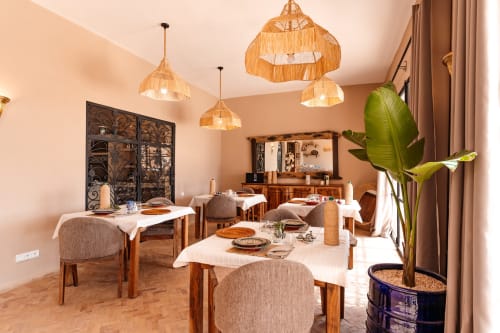 Salle à manger élégante du Riad Arkaya à Essaouira avec luminaires en osier tressé, tables en bois et plantes luxuriantes