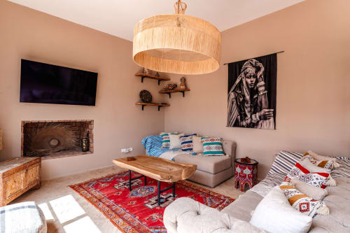 Salon du Riad Arkaya à Essaouira avec luminaire suspendu tressé, tapis rouge, sièges beige et décoration artisanale