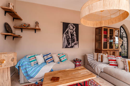 Salon d'hôtes du Riad Arkaya à Essaouira avec deux canapés beiges, coussins marocains colorés, tables en bois et lampe suspension tissée.