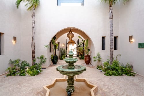 La cour du Riad Arkaya à Essaouira présente une colonnade en arc, une fontaine ornée, des plantes en pots et des palmiers.