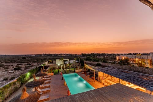 Hôtel Riad Arkaya à Essaouira au coucher de soleil avec piscine turquoise, terrasse en bois, transats et vue sur le paysage désertique.