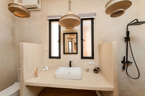 Salle de bains moderne au Riad Arkaya d'Essaouira avec vasque blanche, luminaires suspendus tressés, mitigeur noir et jardin