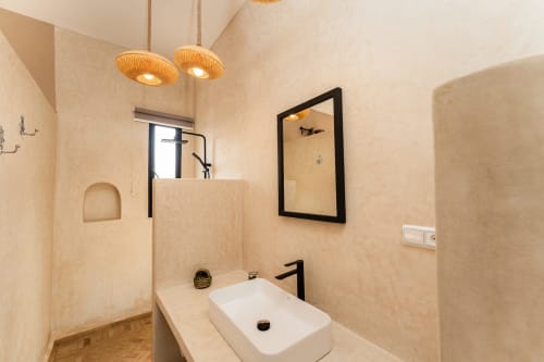 Salle de bain moderne du Riad Arkaya à Essaouira avec lavabo blanc, robinetterie noire, lampes suspendues tissées et douche vitrée