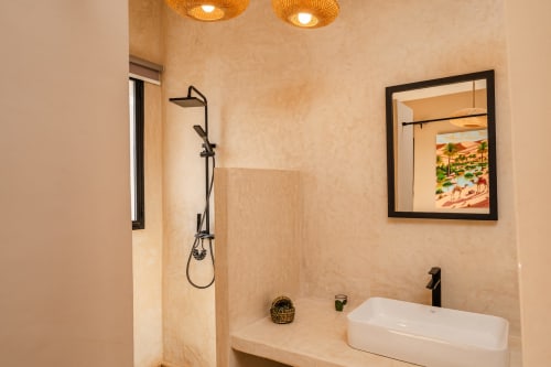 Salle de bains moderne au Riad Arkaya d'Essaouira avec murs beiges, vasque blanche, robinetterie noire et douche pluie