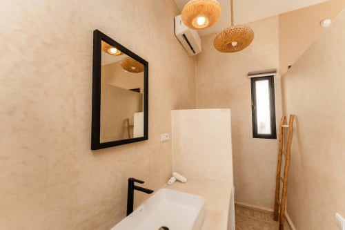 Salle de bains moderne au Riad Arkaya à Essaouira : miroir à cadre noir, luminaires suspendus tissés et vasque minimaliste blanche.