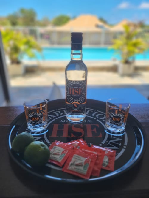 Bouteille de rhum blanc HSE avec verres et citrons verts sur plateau aux couleurs de la marque, vue sur la piscine de Villa Maloem, Sainte-Anne