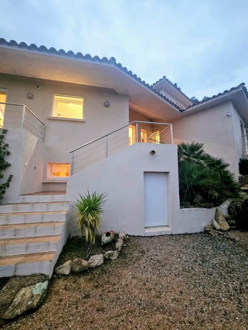 Modern villa entrance at Corsu Di Core in Favone with illuminated windows, terrace, palm trees, and gravel courtyard at dusk