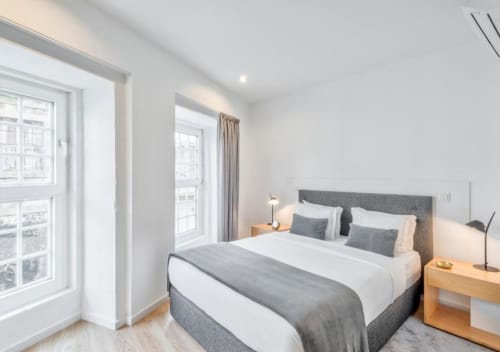 Quarto moderno no Oporto Comfort Dom Hugo com cama estofada cinza, roupa de cama branca, mesinhas de madeira e grandes janelas