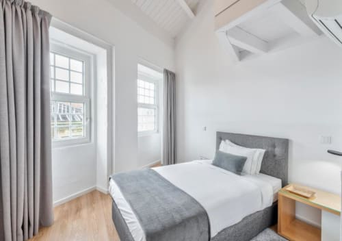 Quarto luminoso do Oporto Comfort Dom Hugo com cama estofada cinzenta, criado-mudo em madeira e janelas amplas com vista para o jardim