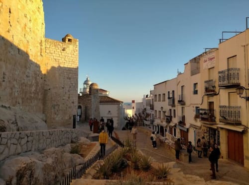 Medieval stone castle and historic buildings in Peñíscola old town with visitors exploring the cobblestone plaza under clear