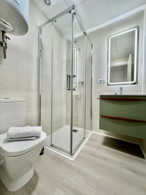 Baño moderno con cabina de ducha cristal, tocador verde, espejo iluminado, inodoro blanco y paredes alicatadas en Architettura