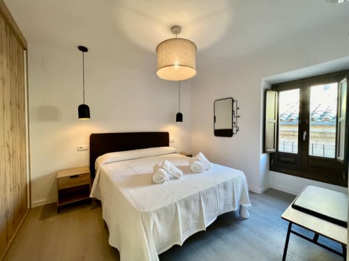 Dormitorio moderno en Architettura Apartamentos de Úbeda con ropa de cama blanca, cabecero oscuro, iluminación colgante y vistas a la ciudad
