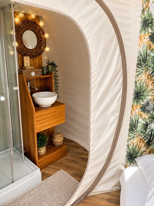 Pod salle de bain luxueux aux formes arrondies à La Biche Au Bois, vanité en bois, miroir rétroéclairé et accents de papier peint tropical.