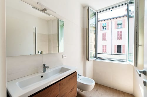 Bagno moderno presso Lago Suites Como con vanity lavabo, specchio, WC e finestra con vista città su edificio storico rosa