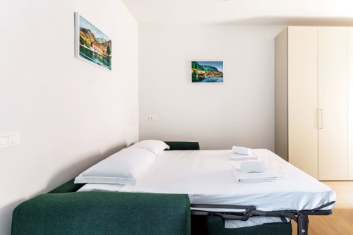 Camera da letto moderna e luminosa a Lago Suites Como con letto bianco, poltrona verde e quadri con veduta lago