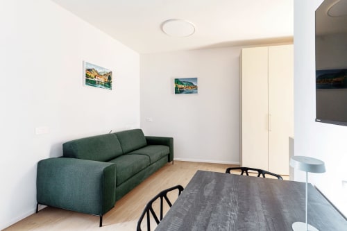 Camera moderna a Lago Suites Como con divano verde, tavolo da pranzo, quadri alle pareti e vista sulla città in TV