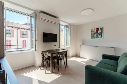 Camera moderna presso Lago Suites Como con vista sulla città, TV, scrivania, divano verde e ampie finestre con vista sull'architettura italiana.