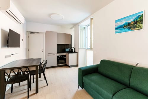 Studio moderno a Lago Suites Como con divano verde, tavolo da pranzo, angolo cottura, TV e opere d'arte lacustre