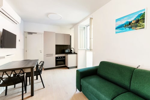 Studio moderno presso Lago Suites Como con divano verde, zona pranzo, cucina, TV e quadri con vista lago