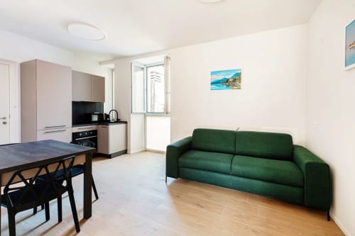 Suite moderna Lago Suites Como con divano verde, tavolo da pranzo, angolo cottura e quadri con vista lago