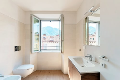 Bagno moderno delle Lago Suites Como con doppio lavabo, wc e finestra con vista sui tetti e le montagne della città