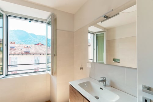 Bagno moderno a Lago Suites Como con finestra vista città, lavabo bianco, specchio e paesaggio montano