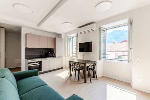 Studio moderno a Lago Suites Como con cucina, zona pranzo, divano teal, TV e vista montagna dal balcone