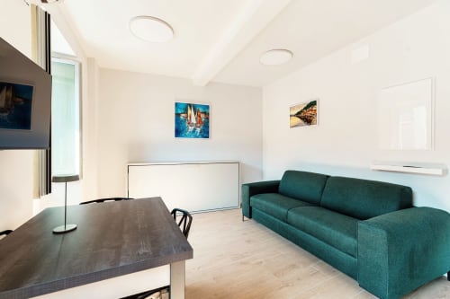 Camera moderna presso Lago Suites Como con divano teal, scrivania, arte murale, TV e vista sulla città