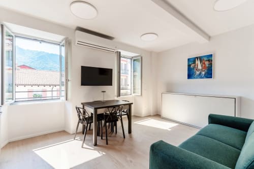 Camera moderna al Lago Suites Como con vista sulla città, tavolo da pranzo, TV schermo piatto, divano verde e grandi finestre panoramiche