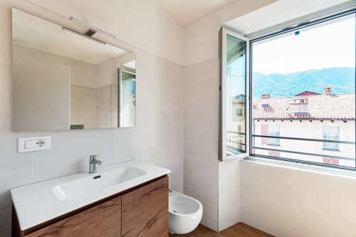 Bagno moderno presso Lago Suites Como con vista sulla città, mobile in legno, specchio e finestra affacciata su tetti e montagne