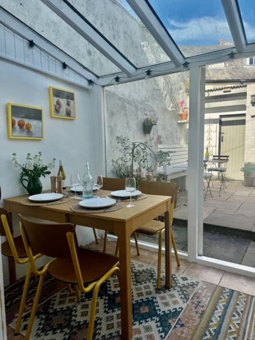 Dining area at 2 Doves Holiday Property in Topsham with wooden table, yellow chairs, and glass roof overlooking garden.
