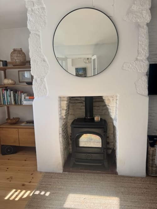 Modern fireplace with black wood stove, circular mirror, and wooden furniture in 2 Doves Holiday Property, Exeter