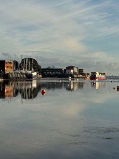 Topsham harbor at dawn with sailboats, red buoys, and waterfront buildings reflected in calm water