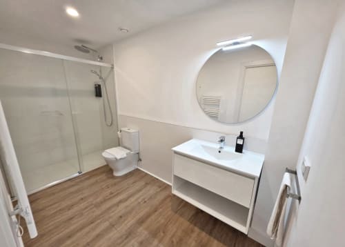 Baño moderno en apartamento BR Silgar Sanxenxo con ducha abierta, espejo redondo, lavamanos blanco y suelos efecto madera
