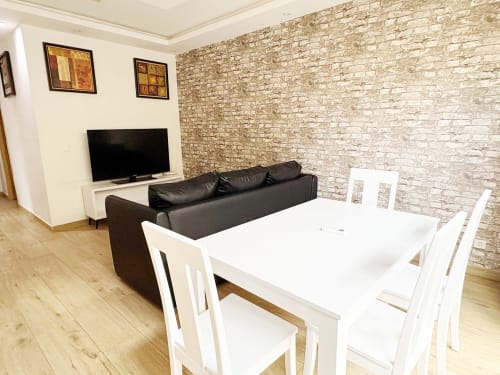 Área moderna de estar y comedor en Dakar con sofá de cuero negro, mesa blanca, pared de ladrillo y televisor