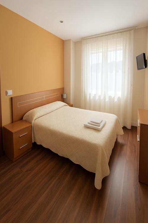 Luminosa habitación doble en la Pensión Gelin con pared naranja, cama de madera, ropa de cama crema y ventana con cortinas translúcidas