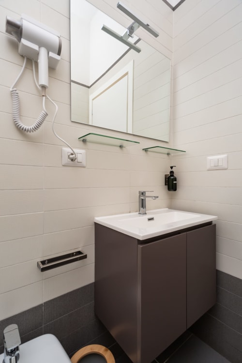 Bagno moderno in Darna Rooms Napoli: mobile scuro, lavabo bianco, specchio, ripiani in vetro, rivestimenti, asciugacapelli