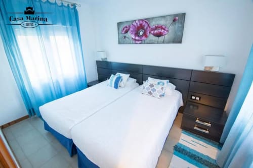 Habitación de Casa Marina Suites con camas gemelas, cabecera oscura, cuadro de flores moradas y cortinas azules en ES