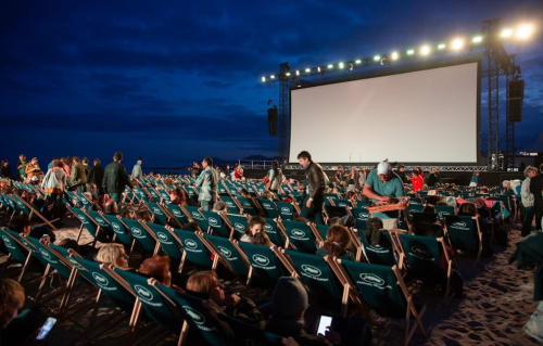 Cine al aire libre en la playa al atardecer con sillas turquesas frente a pantalla gigante, cielo crepuscular, multitud reunida para la noche de cine