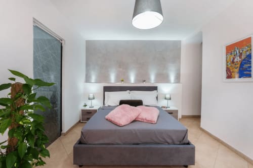 Camera moderna alle Origenes Rooms, Procida con letto grigio, cuscini rosa, pianta e illuminazione contemporanea