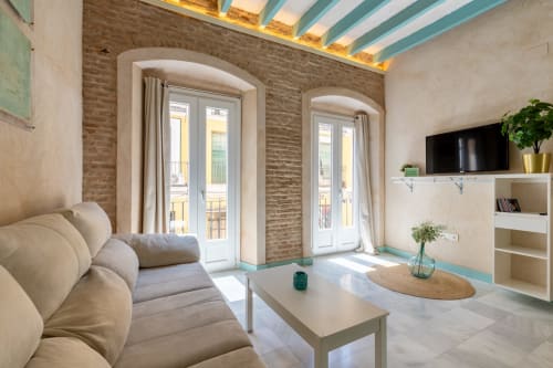 Moderno apartamento luminoso en Sevilla con sofá crema, paredes de ladrillo visto, vigas turquesas y puertas de cristal al balcón
